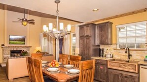 A dining area with a wooden table and chairs, a chandelier, a fireplace, and a kitchen with dark wooden cabinets and a window. - Rustic 2BR Condo w/ Views & Full Kitchen (Pigeon Forge)