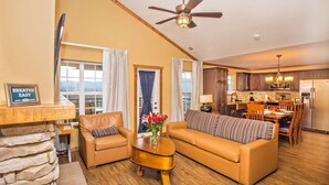 A spacious living room with a sofa, two armchairs, a coffee table, and a stone fireplace. A kitchen is visible in the background with wooden cabinets and a dining area. - Rustic 2BR Condo w/ Views & Full Kitchen (Pigeon Forge)