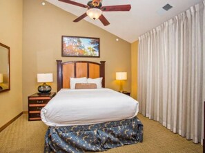 Una habitación de hotel con cama, ventilador de techo, un cuadro en la pared y ventana con cortinas.