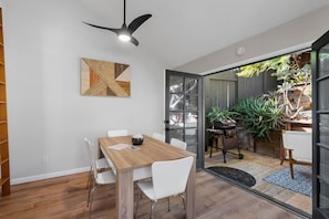 Un espace repas moderne avec une table en bois et des chaises blanches, un ventilateur de plafond et une vue sur un jardin, visible à travers des portes vitrées.