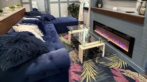 Ein modernes Wohnzimmer mit einem blauen Ecksofa, einem Glastisch, einem Kamin und einem wandmontierten Fernseher.