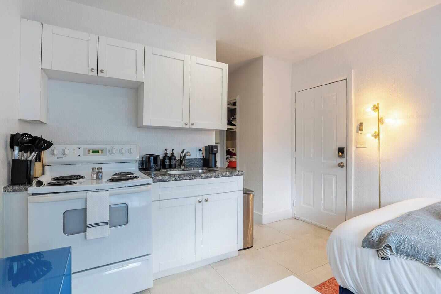 Une cuisine moderne avec des armoires blanches, une cuisinière et un évier. On y trouve un lit avec une couverture grise et un petit tapis.