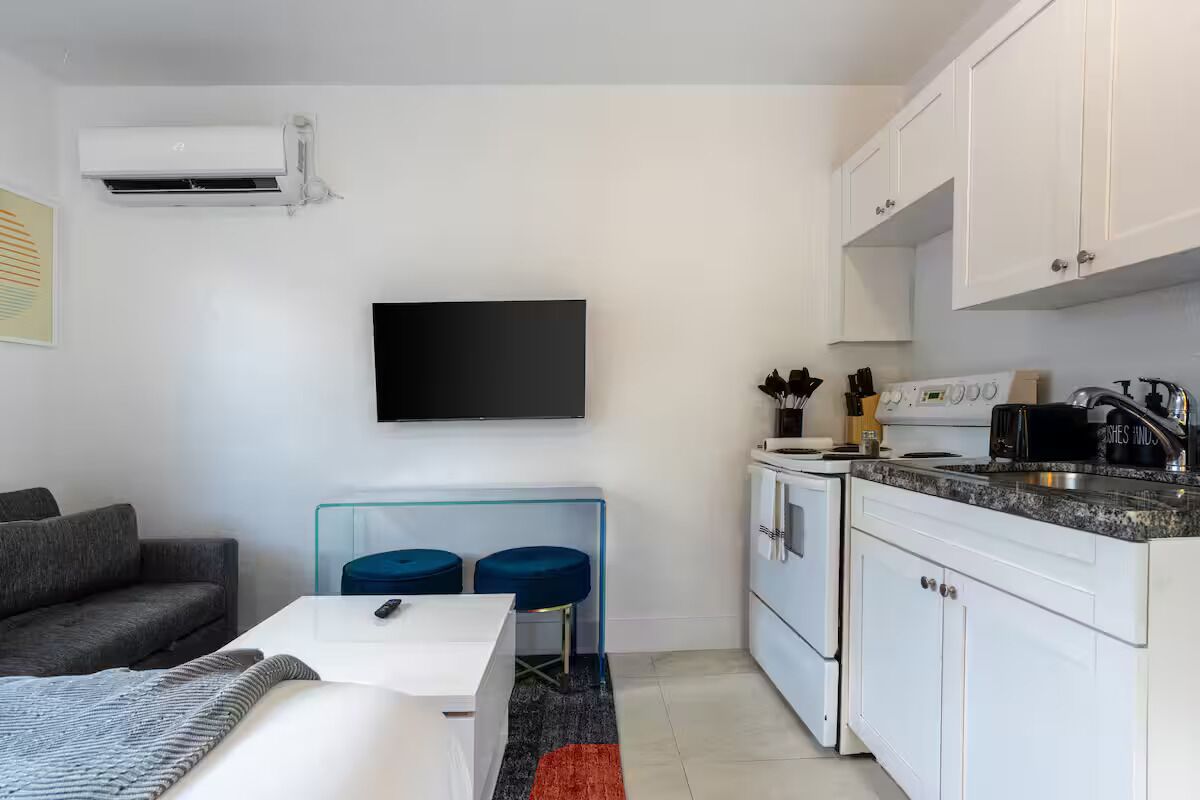 A compact living space with a sofa, a small dining table, a TV mounted on the wall, a kitchenette with a stove and sink, and a glass partition separating the dining area from the living space.