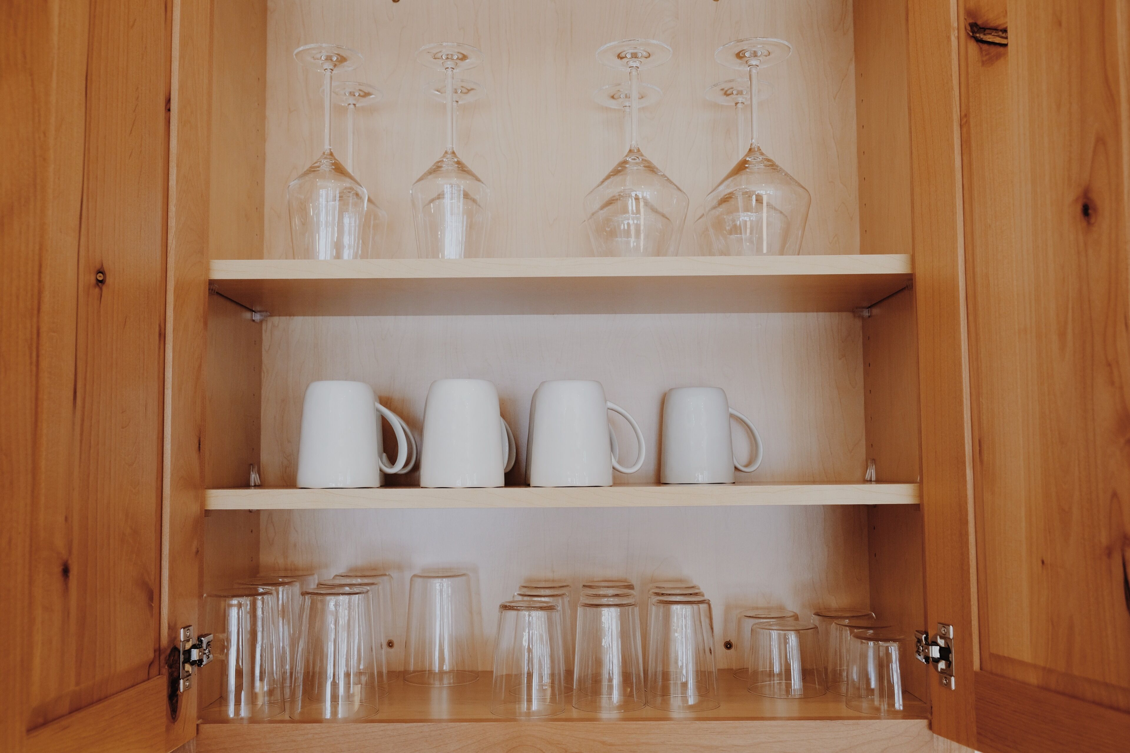 Een houten kast met drie planken. Bovenop staan wijnglazen, in het midden witte mokken en onderin heldere drinkglazen.