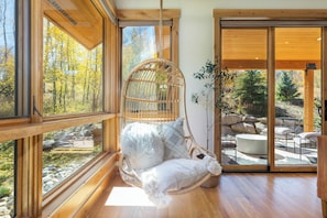 Ein Hängekorbstuhl mit flauschigem Kissen und Polster, platziert bei einem großen Fenster mit Blick auf einen Wald und eine Außenbereich-Sitzgelegenheit.