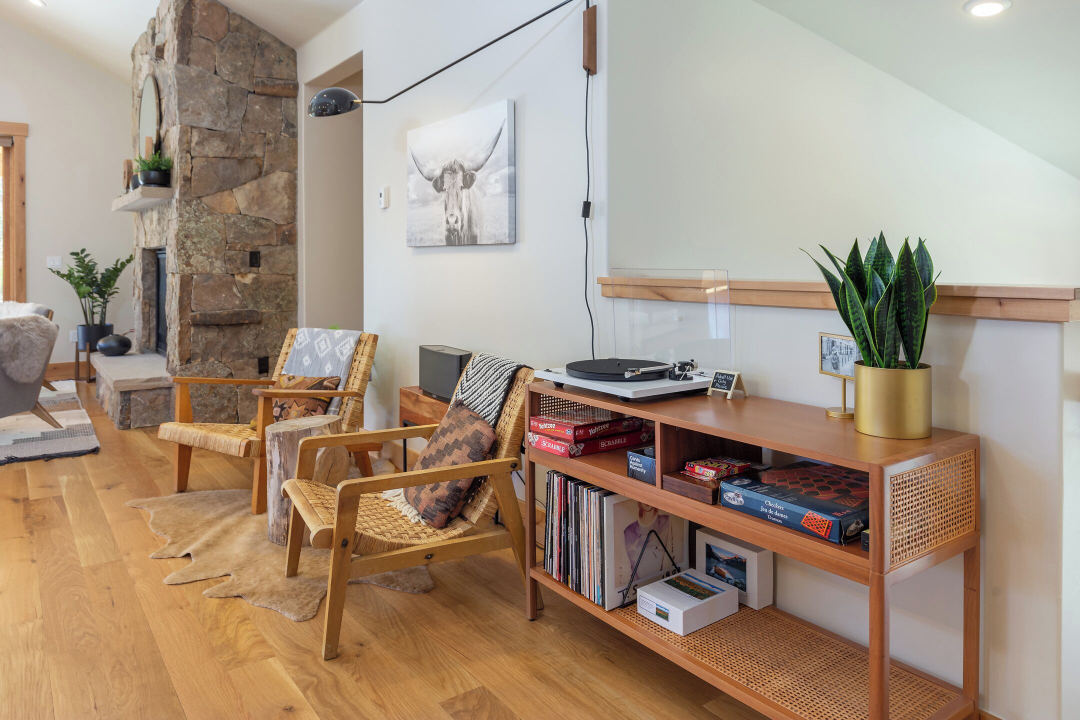 Una sala de estar acogedora con chimenea de piedra, sillas de madera, una estantería y un cuadro colgado de una vaca.