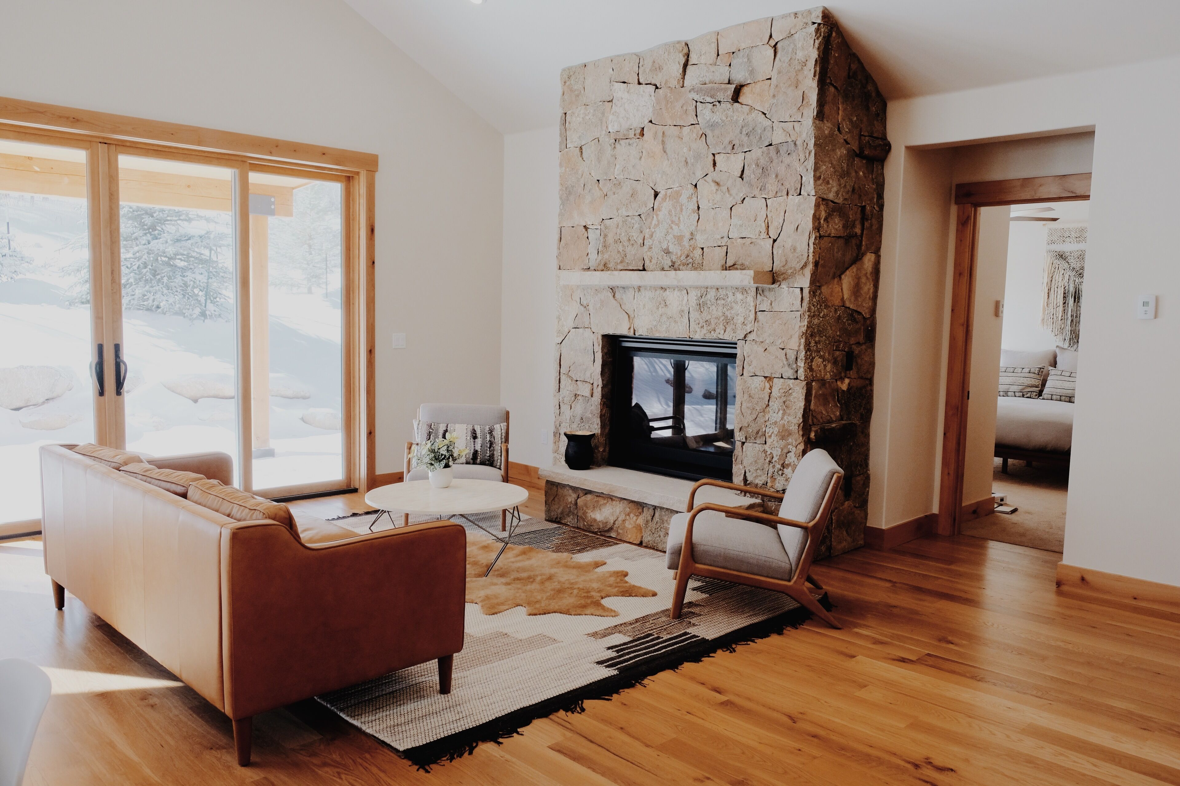 A modern living room with a stone fireplace, wooden flooring, and a large window overlooking a snowy landscape.