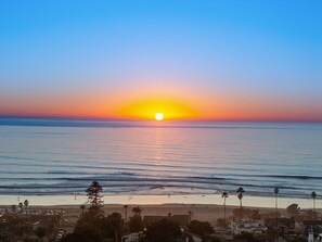 Un atardecer sobre una playa con olas, palmeras y edificios al frente.