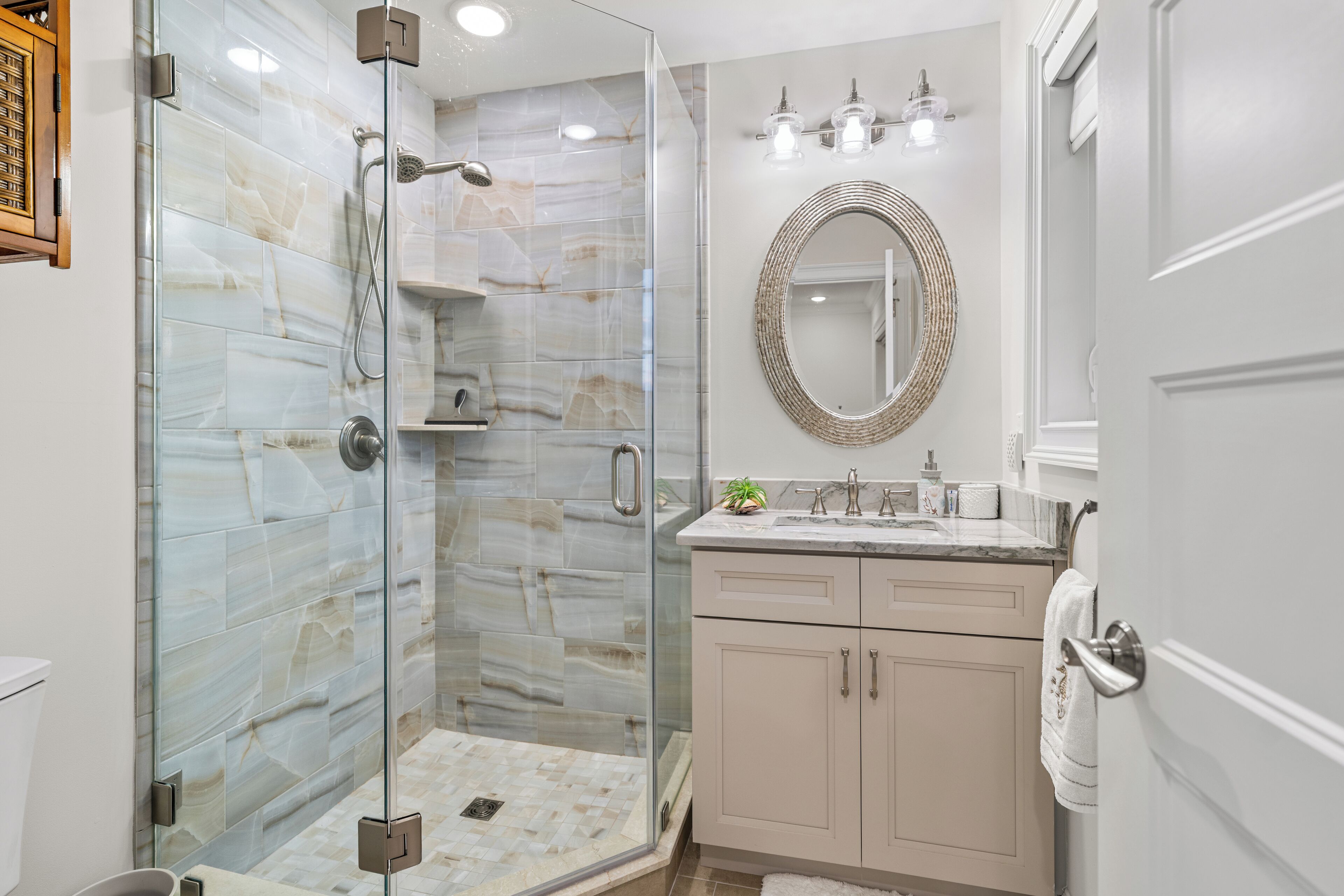 A modern bathroom with a glass-enclosed shower, marble tiles, a sink with a mirror, and beige cabinets.