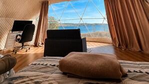 A room with a geometric ceiling design, a bed, a desk with a chair, and a view of the outdoors. - Baddeck Domes (Baddeck)