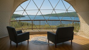 A tent with a large window offering a view of a lake and hills. Inside, there are two chairs and a round rug. - Baddeck Domes (Baddeck)