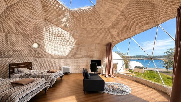 A spacious room with a high ceiling, two beds, a desk, and a large window offering a view of the sea. - Baddeck Domes (Baddeck)