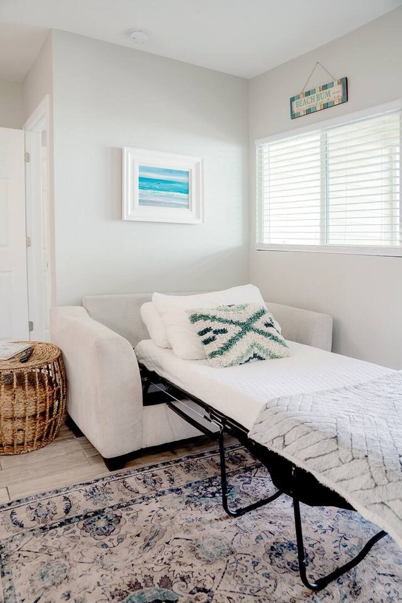 A minimalist bedroom with a bed, a woven basket, a framed picture, and a window with blinds.