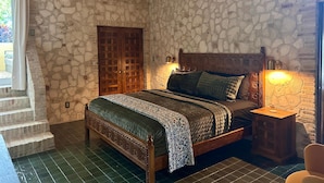 A bedroom with a stone wall, a wooden bed with a headboard, a nightstand, a window with curtains, and an air conditioning unit. - Treehouse - The Canopy (Christiansted)
