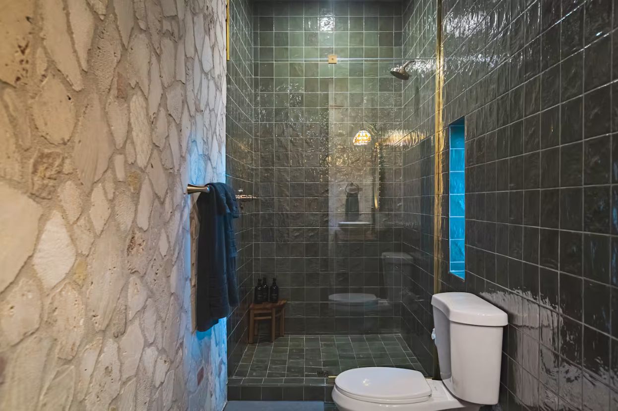 Ein Badezimmer mit Steinwand, einer gefliesten Duschkabine, einer Toilette und einem kleinen hölzernen Hocker mit Flaschen.