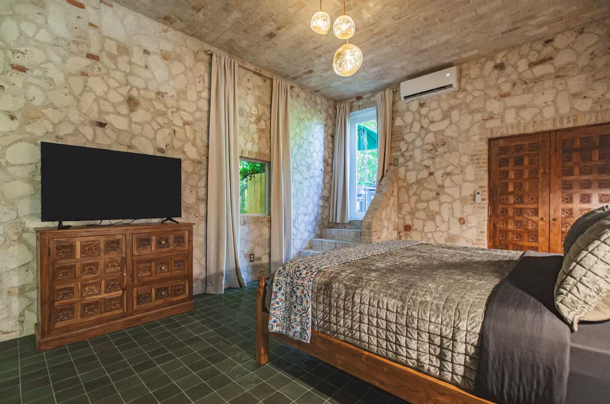 Un dormitorio con pared de piedra, cama de madera, televisor sobre un mueble y una escalera que lleva a un balcón.