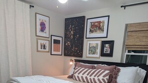 Un lit bien fait, agrémenté d’un coussin à motifs, un mur orné de tableaux encadrés et une fenêtre avec un store.