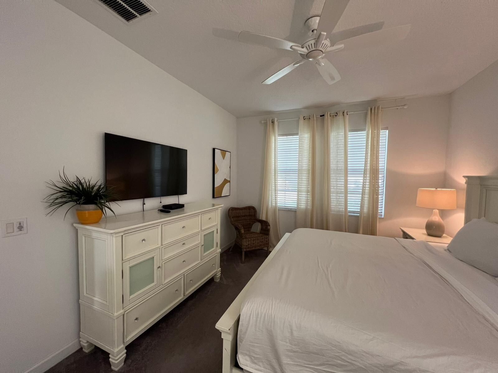 A bedroom with a bed, a TV, a ceiling fan, a dresser, a chair, and a window with curtains.