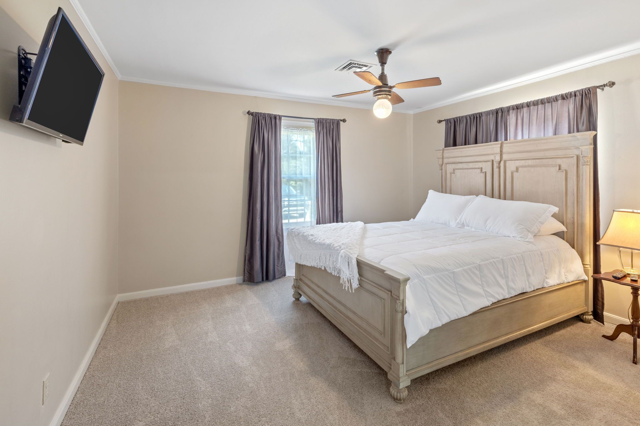 Une chambre à coucher avec un lit, un ventilateur de plafond, un téléviseur fixé au mur et une fenêtre avec des rideaux.
