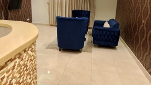 A room with a curved reception desk, two blue armchairs, a patterned wall, and a large window with curtains. - Diyra Hira (Jeddah)