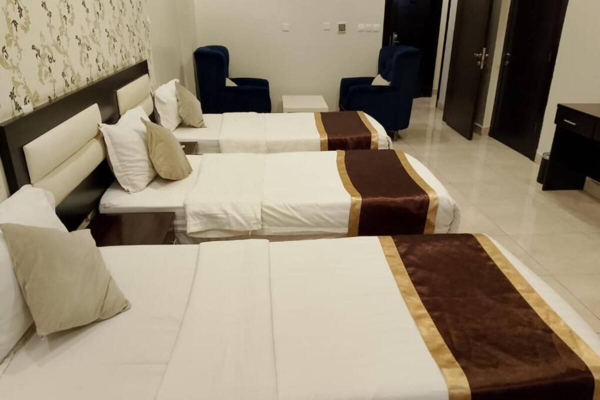 Une chambre d’hôtel avec deux lits, chacun recouvert d’une courtepointe blanche agrémentée d’un motif marron. On y trouve deux chaises bleues et une petite table blanche.