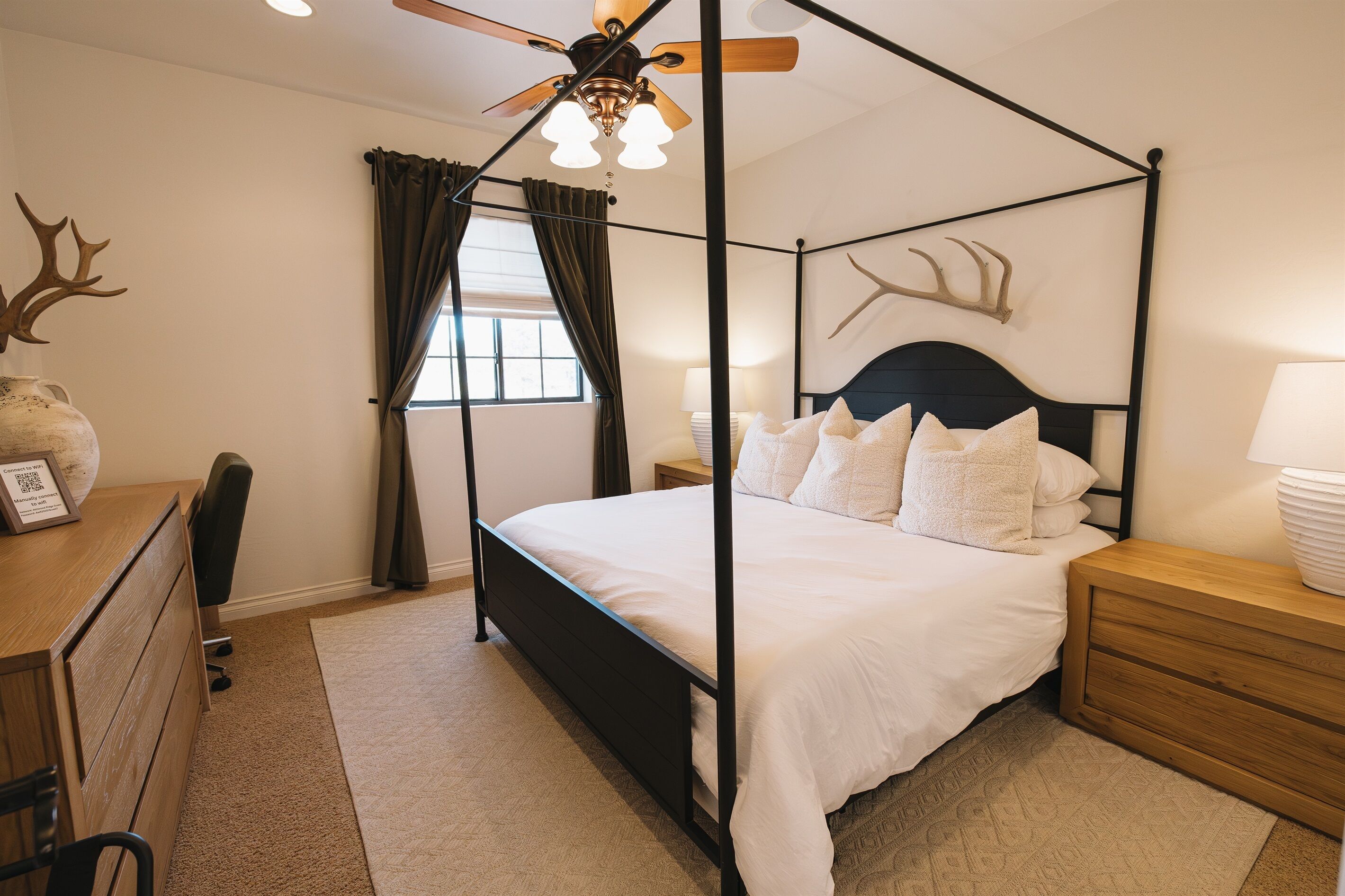 A four-poster bed with white bedding, a wooden nightstand, a desk with a chair, a ceiling fan, and a window with curtains.