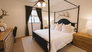 A four-poster bed with white bedding, a wooden nightstand, a desk with a chair, a ceiling fan, and a window with curtains.