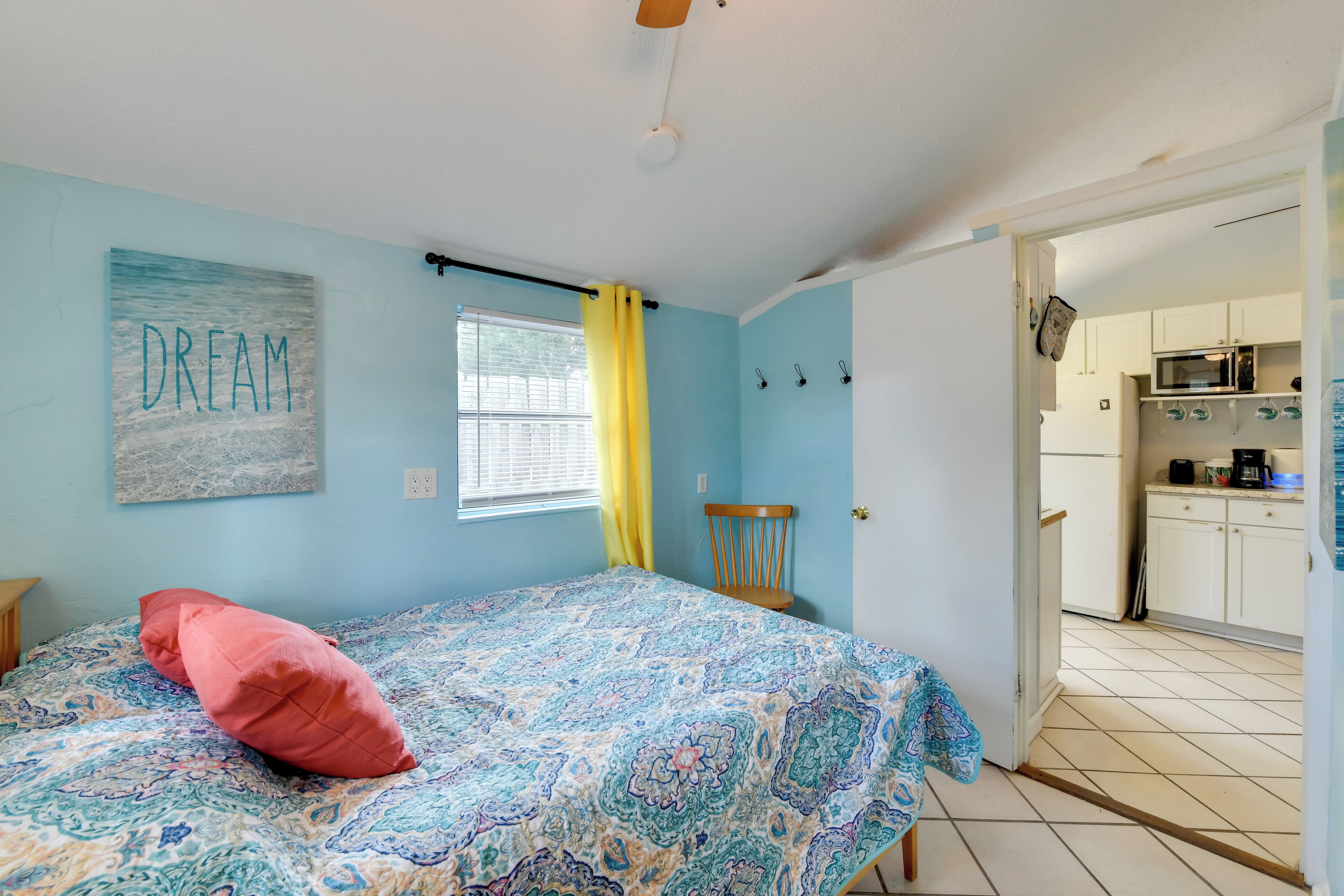 A bedroom with a bed, a window with yellow curtains, a chair, and a kitchen visible through an open door.