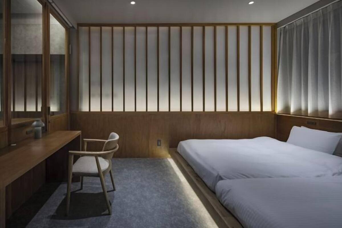 A hotel room with a wooden desk, two beds, a chair, and a large window with blinds.