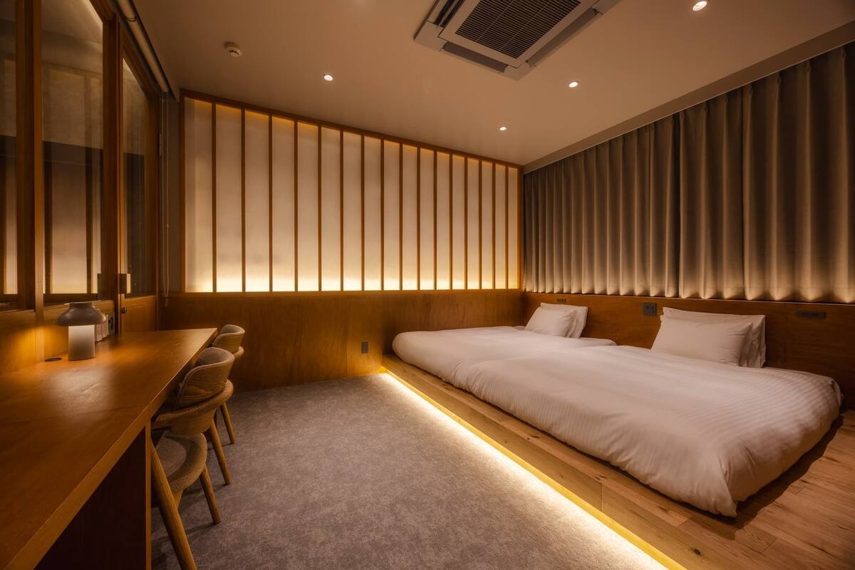 A modern hotel room with a wooden desk, a single bed, and a large window with vertical blinds.
