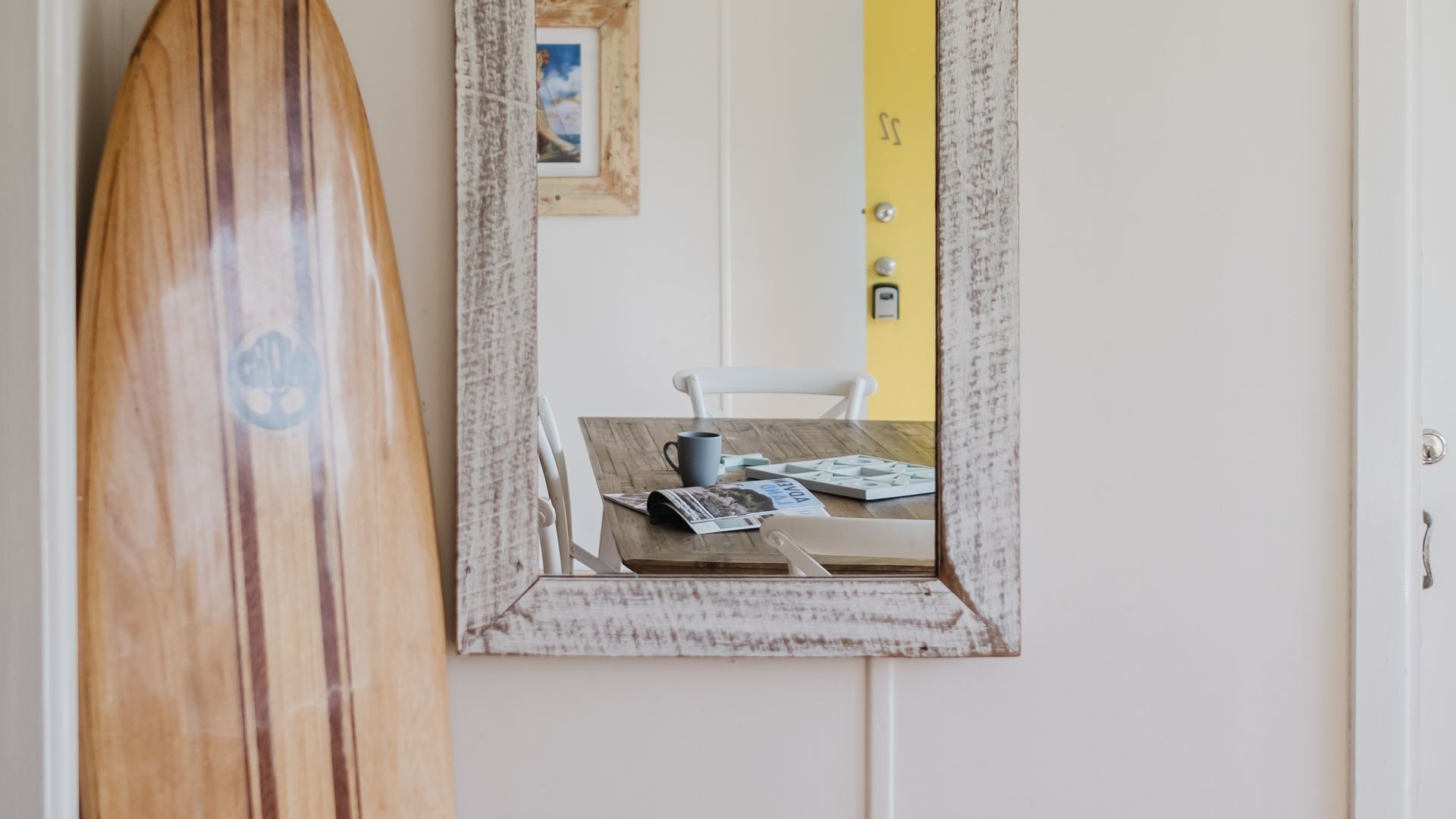 Ein Surfbrett lehnt an einer Wand, ein Spiegel spiegelt einen Tisch mit einer Tasse und Zeitschriften wider, und eine gelbe Tür mit einem kleinen Bilderrahmen.