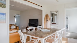 Un comedor con una mesa de madera y sillas blancas, un televisor y una tabla de surf en la pared.