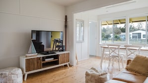 Un salón con televisor, mueble de madera para equipo audiovisual, muebles blancos y un ventanal con vistas al exterior.