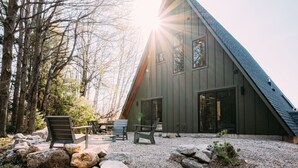 A modern, triangular wooden cabin with a large glass door, surrounded by a gravel area with rocks and a few wooden chairs. - New A-Frame in wine country (Lake Leelanau)