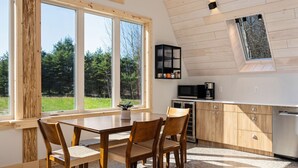 A modern kitchen with wooden cabinets, a dining table with chairs, and a large window overlooking a green landscape. - New A-Frame in wine country (Lake Leelanau)