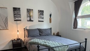 Un dormitorio con cama, mesitas de noche, decoración en la pared, una ventana con cortinas y una lámpara.