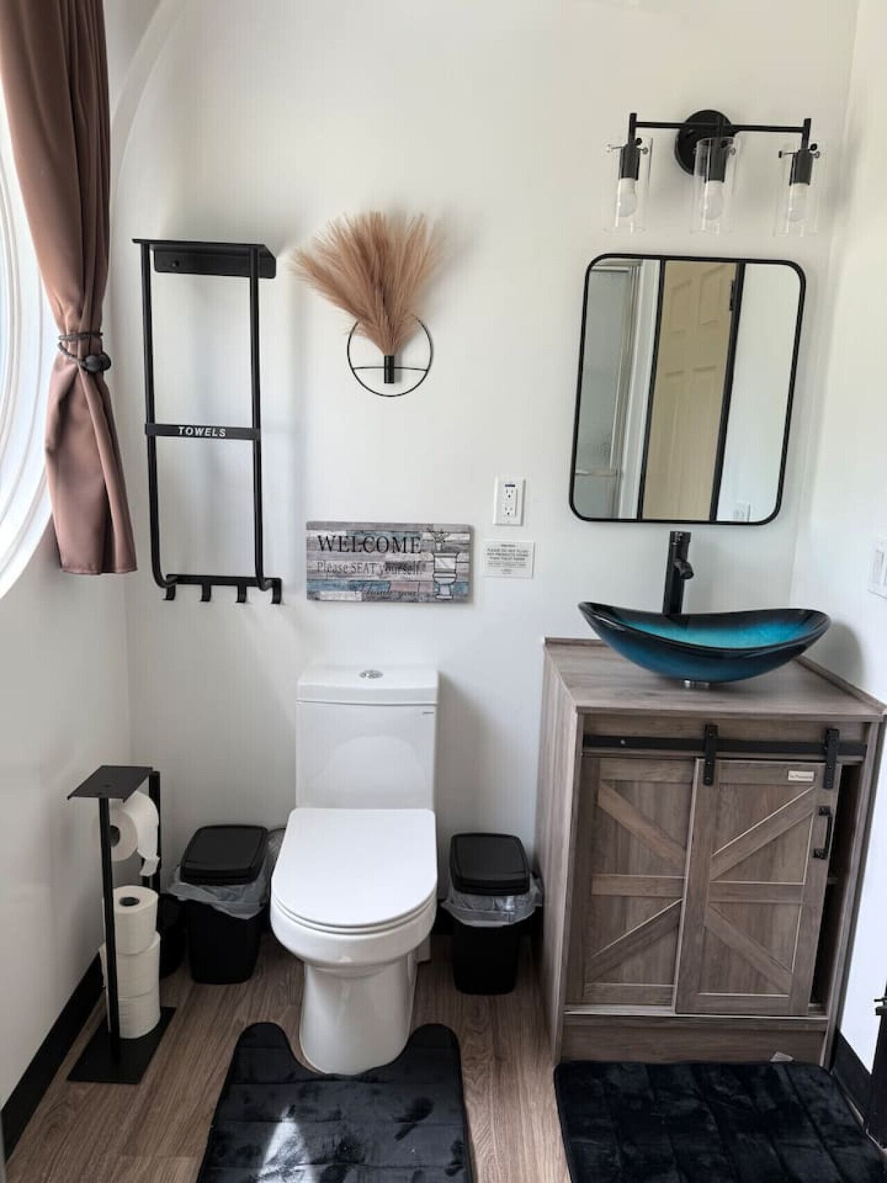 Une petite salle de bain comprenant des toilettes, deux poubelles, un porte-serviettes, un panneau d’accueil, un miroir et un vase décoratif.