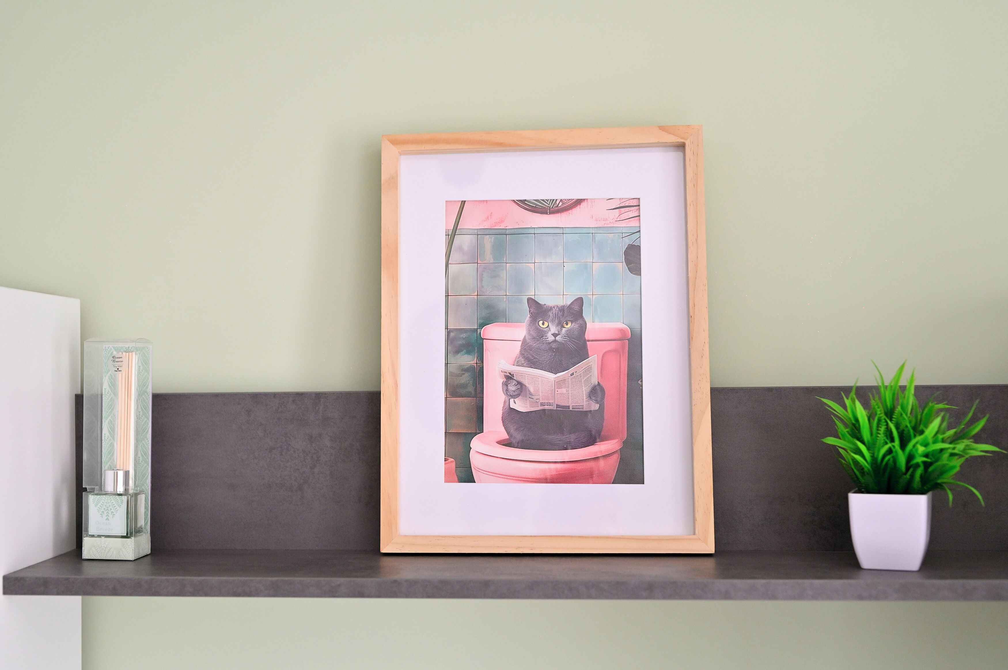 A framed artwork of a cat reading on a toilet, placed on a shelf with a potted plant beside it.
