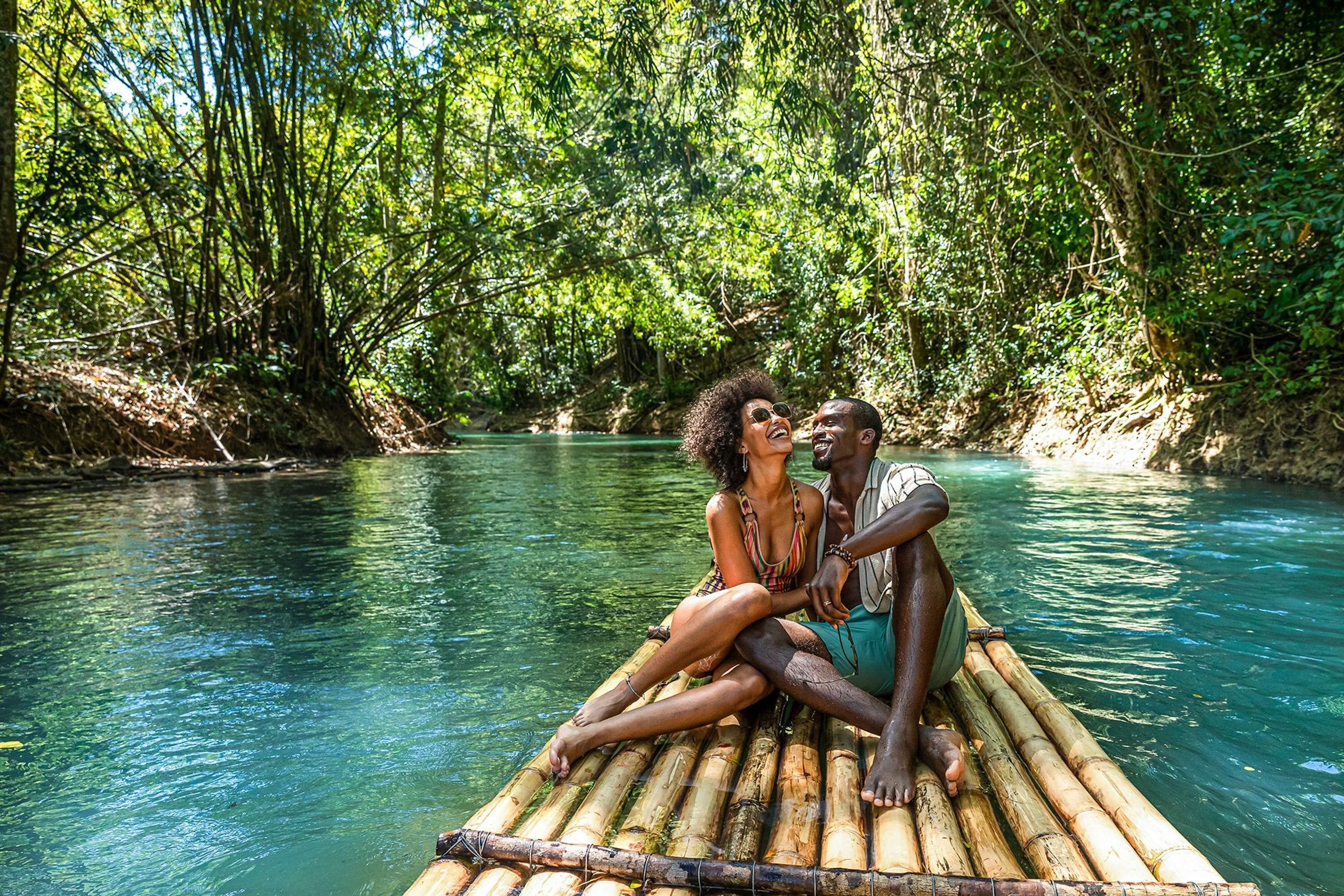 A couple sitting on a bamboo raft in a river surrounded by dense greenery.