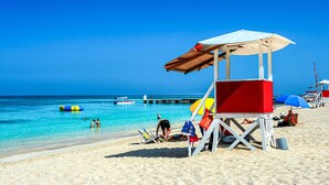 A beach with a lifeguard tower, clear blue water, and a pier in the background. - Luxury Oceanfront Residence Condo at Soleil Residences (Montego Bay)