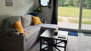 Een moderne woonkamer met een grijze bank, gele en groene kussens, een salontafel en uitzicht op een groene buitenruimte door een schuifpui.