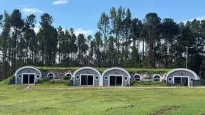 A row of four small, semi-circular buildings with large doors, set in a grassy field with a forest in the background. - Private Earth Home Retreat, mini goats, fire pits and more - unit A (Littleton)