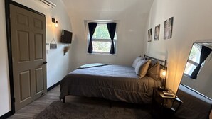 A bedroom with a bed, bedside lamp, window with curtains, wall-mounted TV, and a door. - Private Earth Home Retreat, mini goats, fire pits and more - unit A (Littleton)