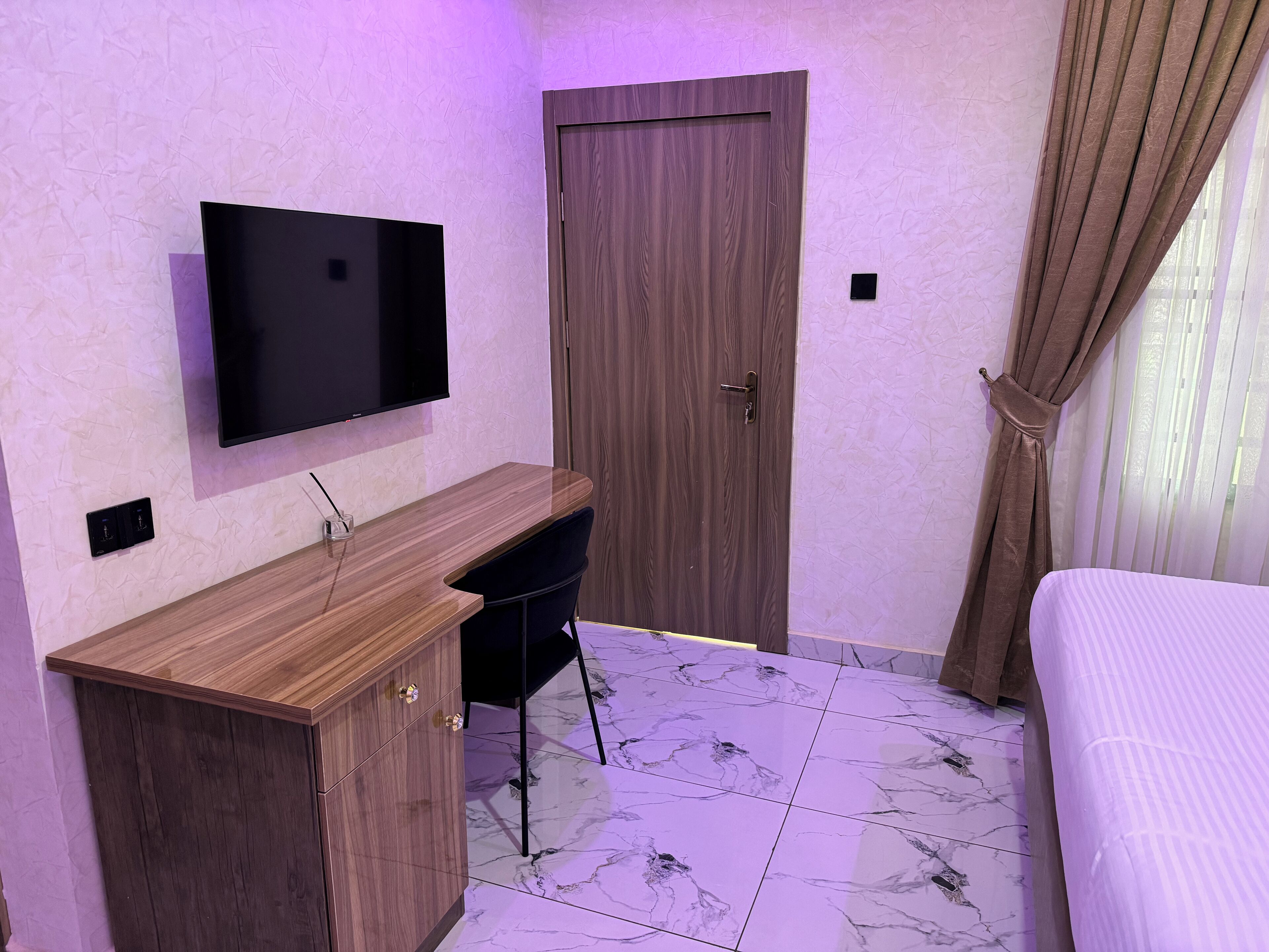 Un dormitorio con cama, escritorio, televisión, puerta y ventana con cortinas.