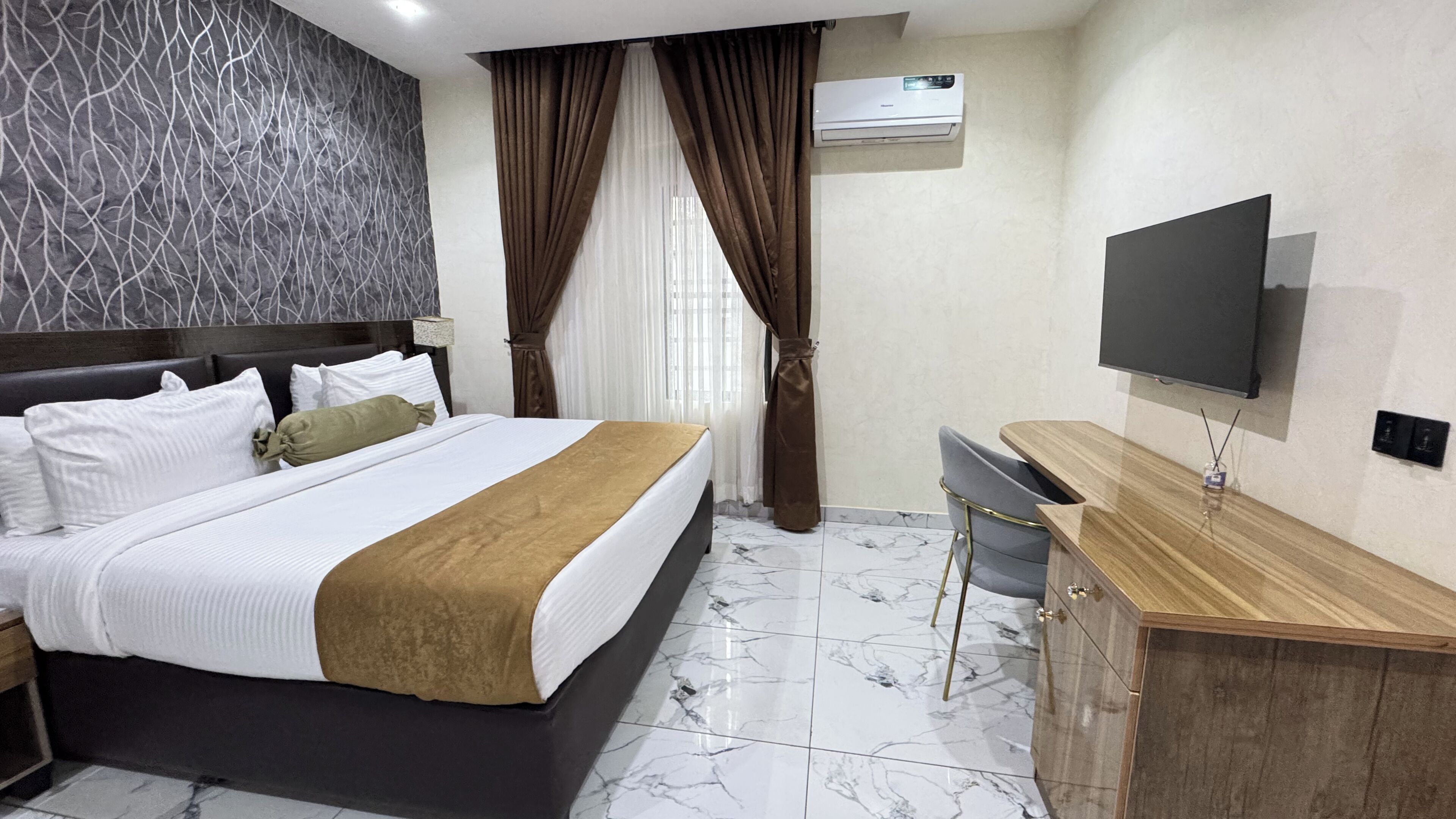 A hotel room with a bed, a desk, a TV, and an air conditioning unit.