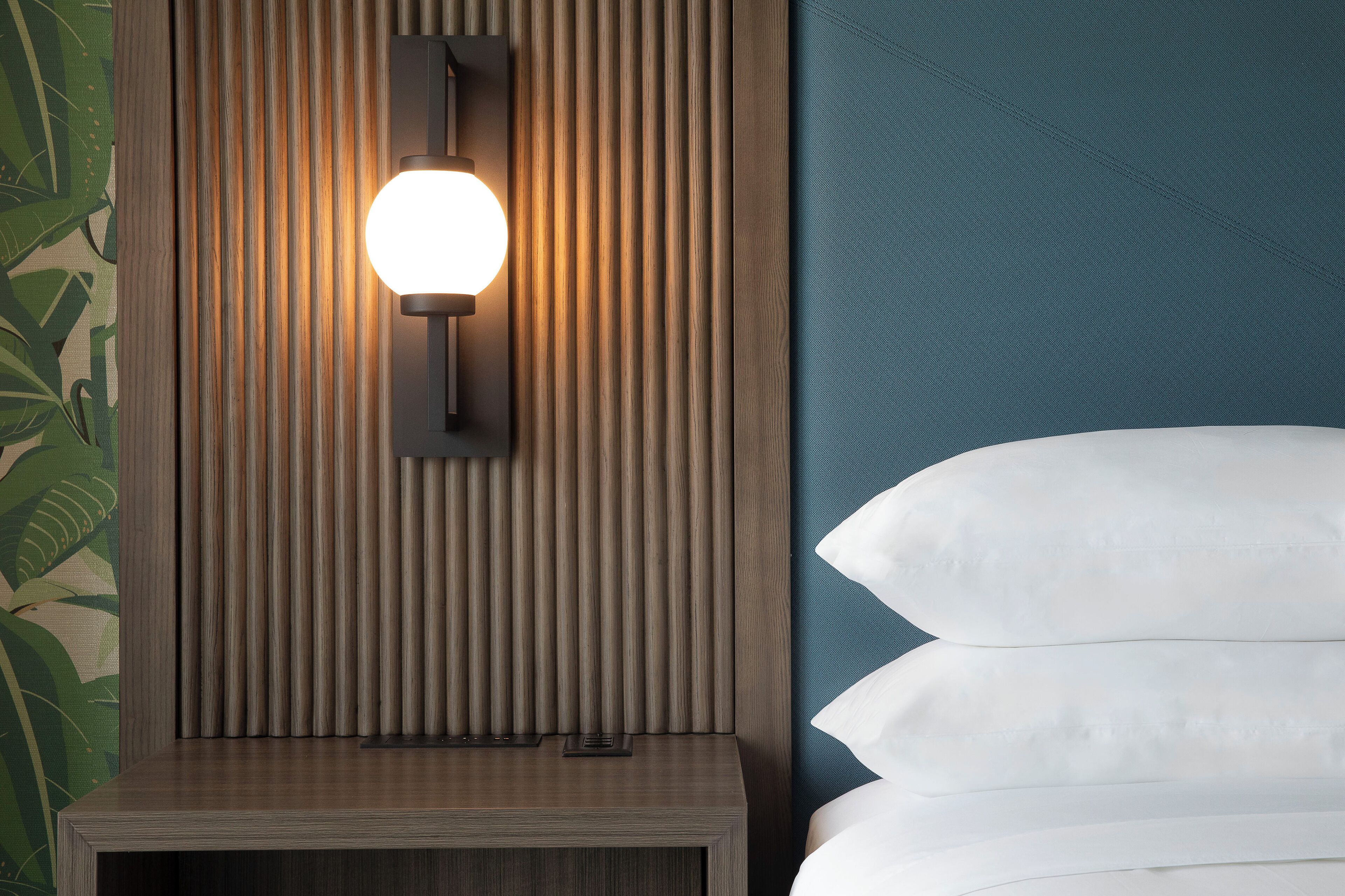 A neatly made bed with white pillows and a wooden bedside table with a wall-mounted light fixture.