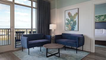 Quarto de hotel com dois sofás azuis, uma mesa de centro redonda, um abajur e uma janela grande com cortinas.