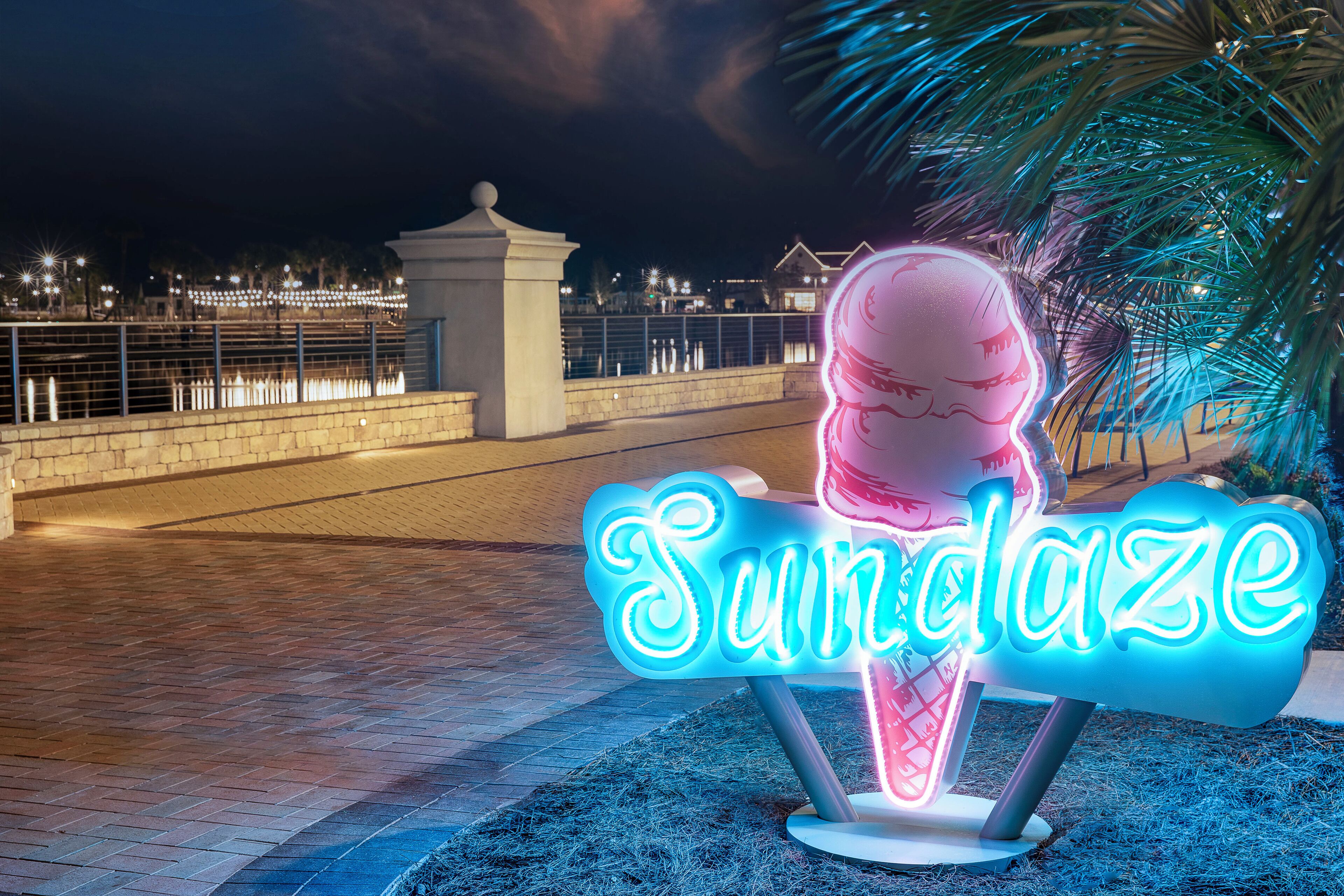 A neon sign with the word "Sundaze" in a stylized font, placed on a brick pathway with a palm tree in the background.