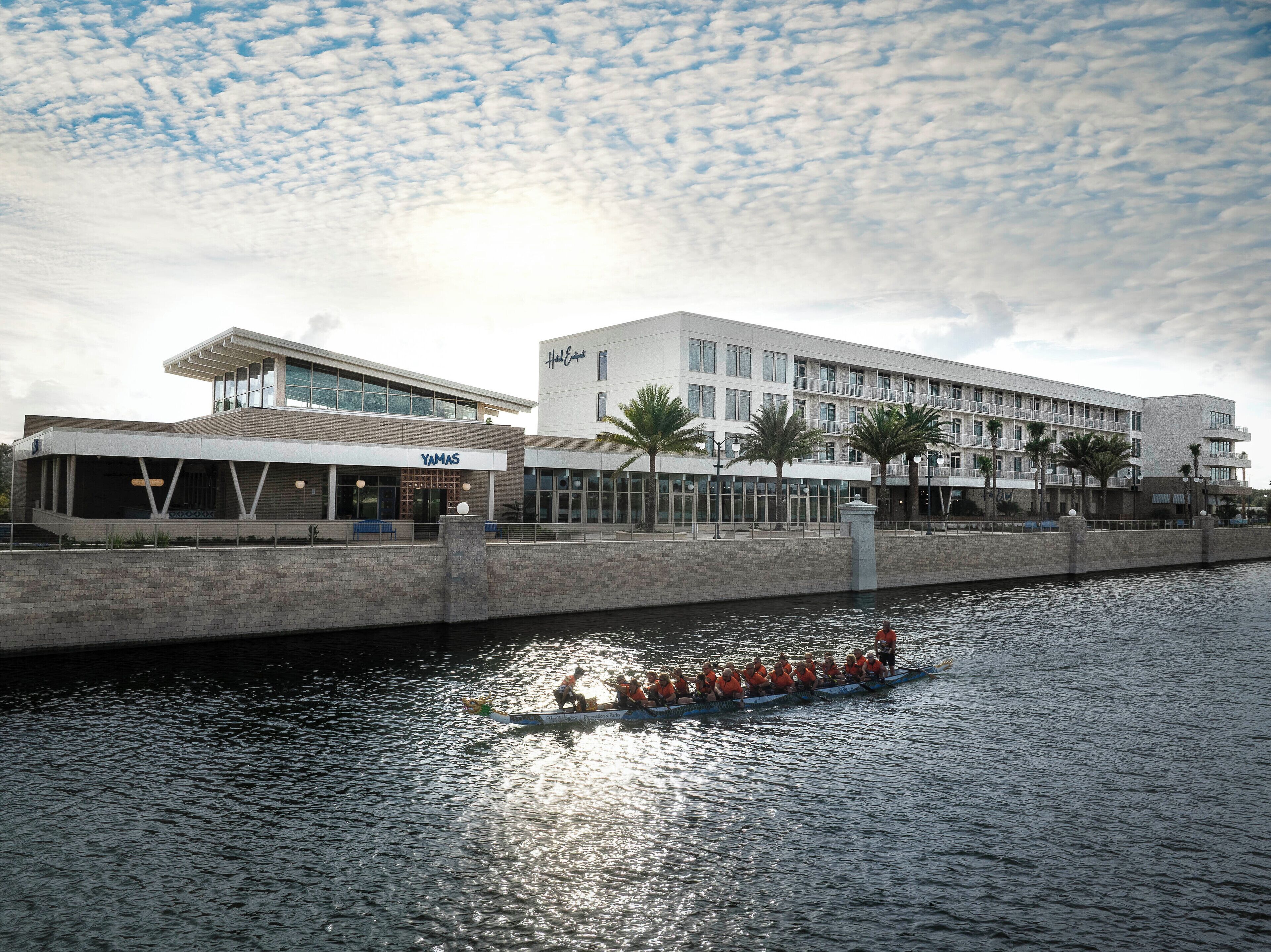 A waterfront hotel with a modern design, featuring a YAMAS restaurant, a pool area, and a view of the water.