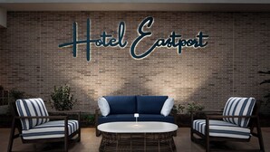 Un espace détente avec un canapé bleu et deux fauteuils à rayures, une table basse ronde et une enseigne indiquant « Hôtel Eastport » sur un mur de briques.